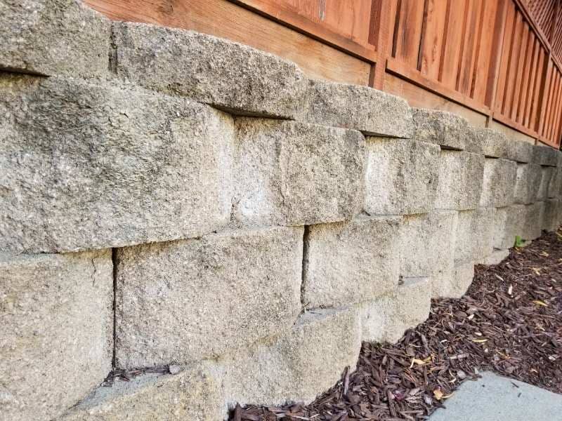 Surveyor measuring ground elevations beside a residential retaining wall where runoff drains through weep holes into a lower yard.